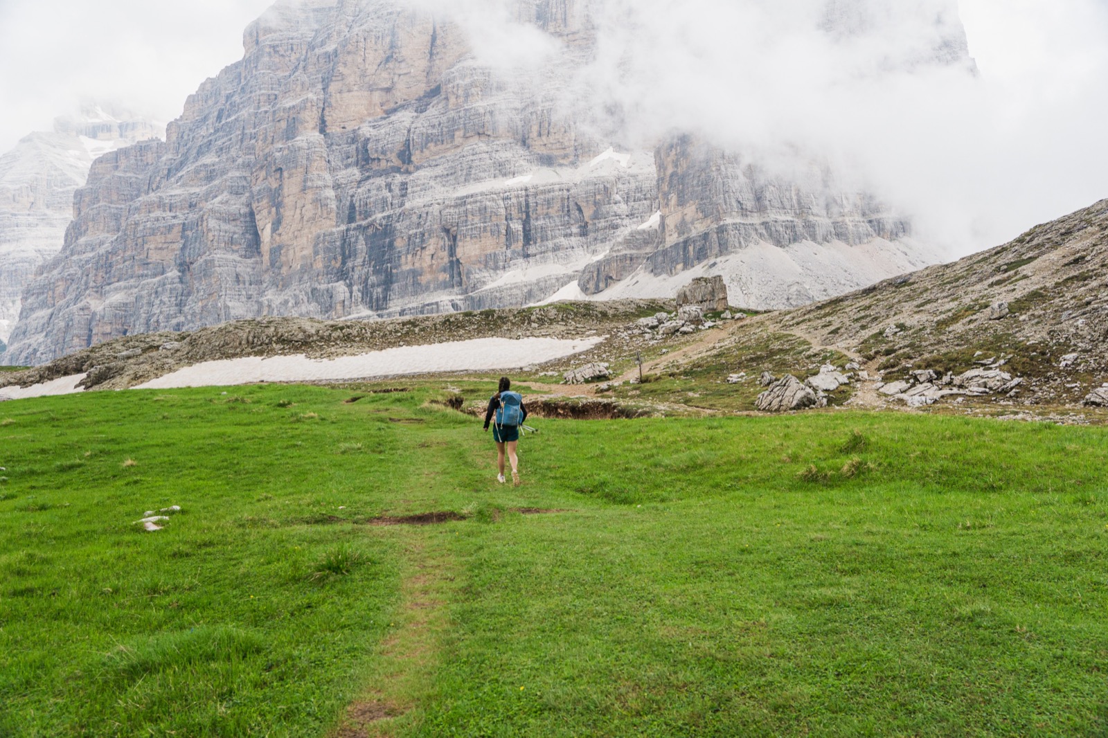 Runner crossing a green meadow toward cloud-wrapped Dolomite cliffs