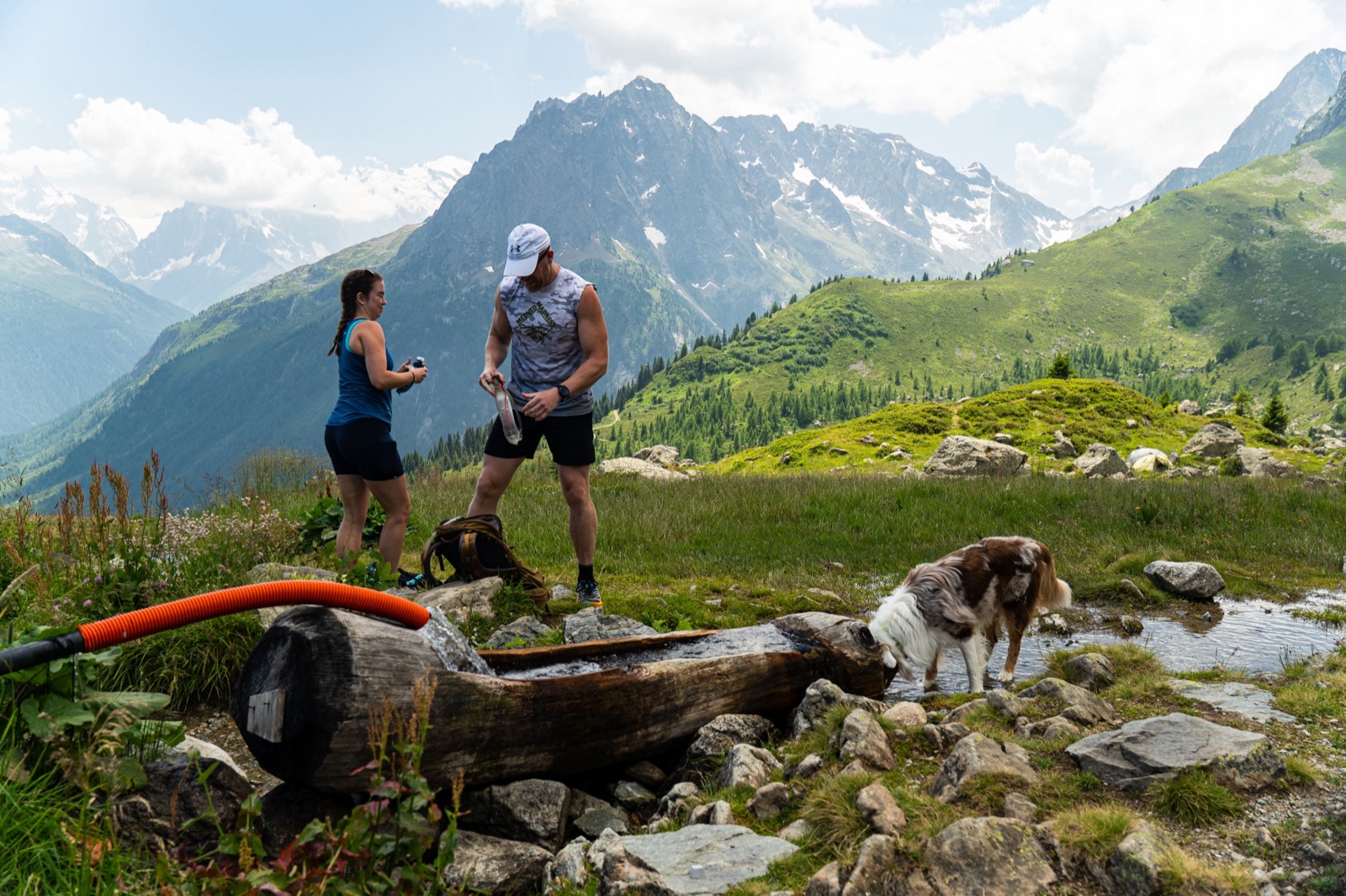 Two runners by a wooden water trough on an alpine pasture, with a dog nearby