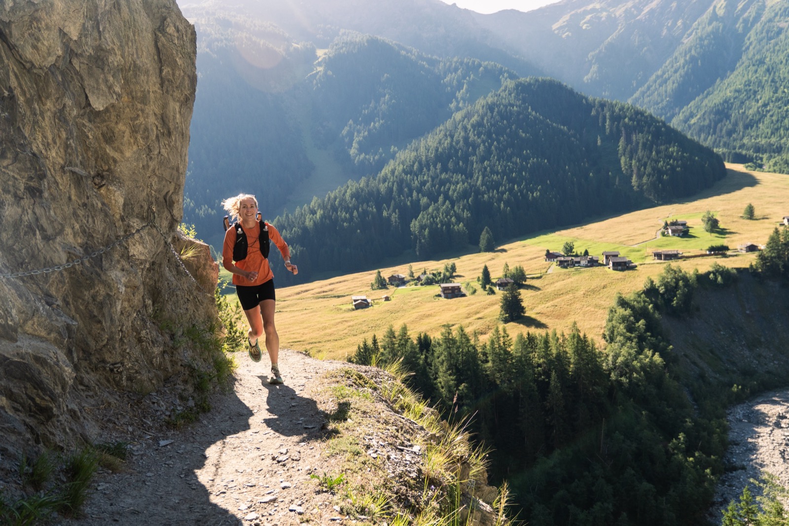 Runner on a cliffside trail above a sunlit valley dotted with chalets