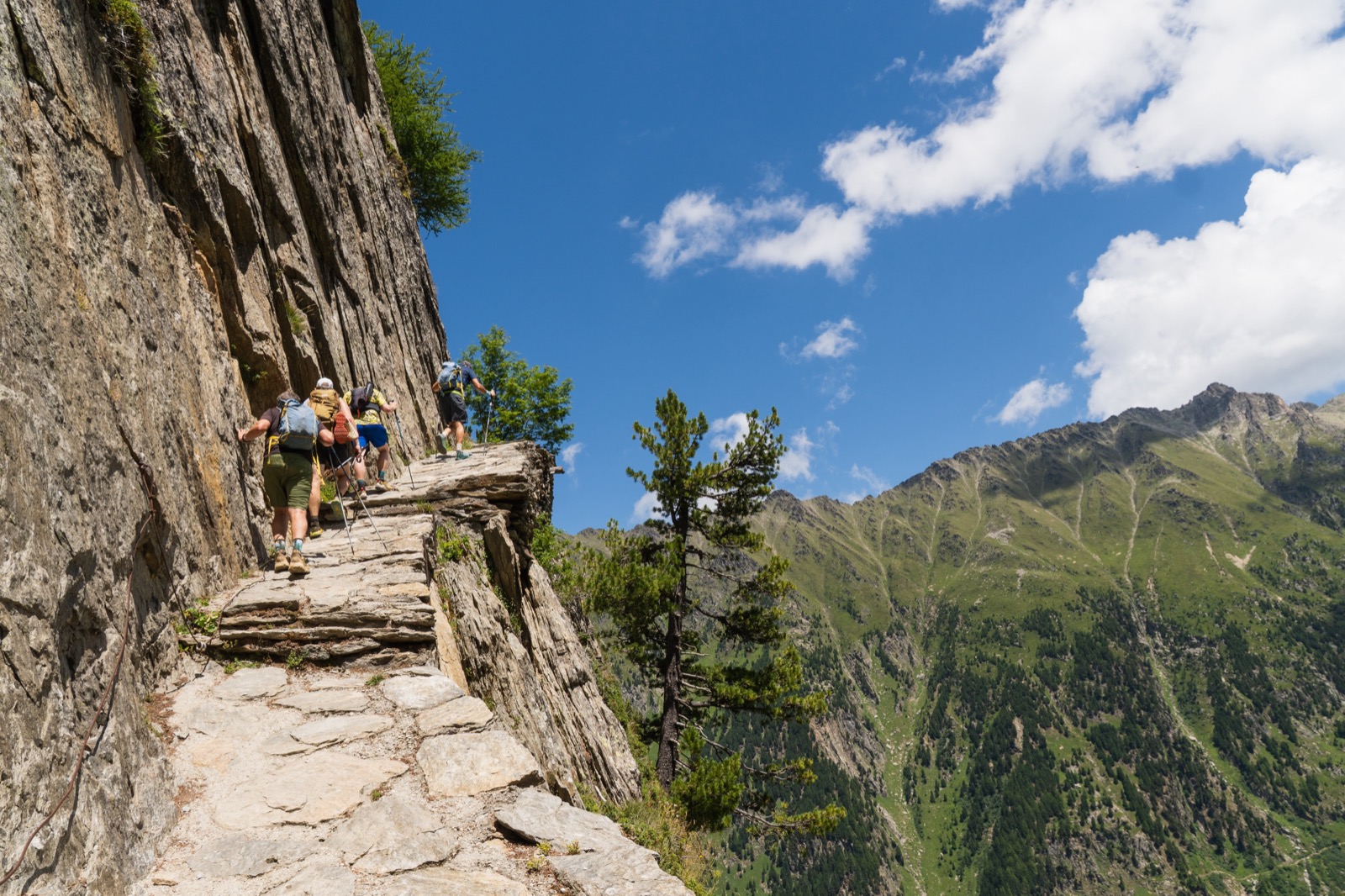 Runners on a stone-paved trail cut into a cliff above a green valley