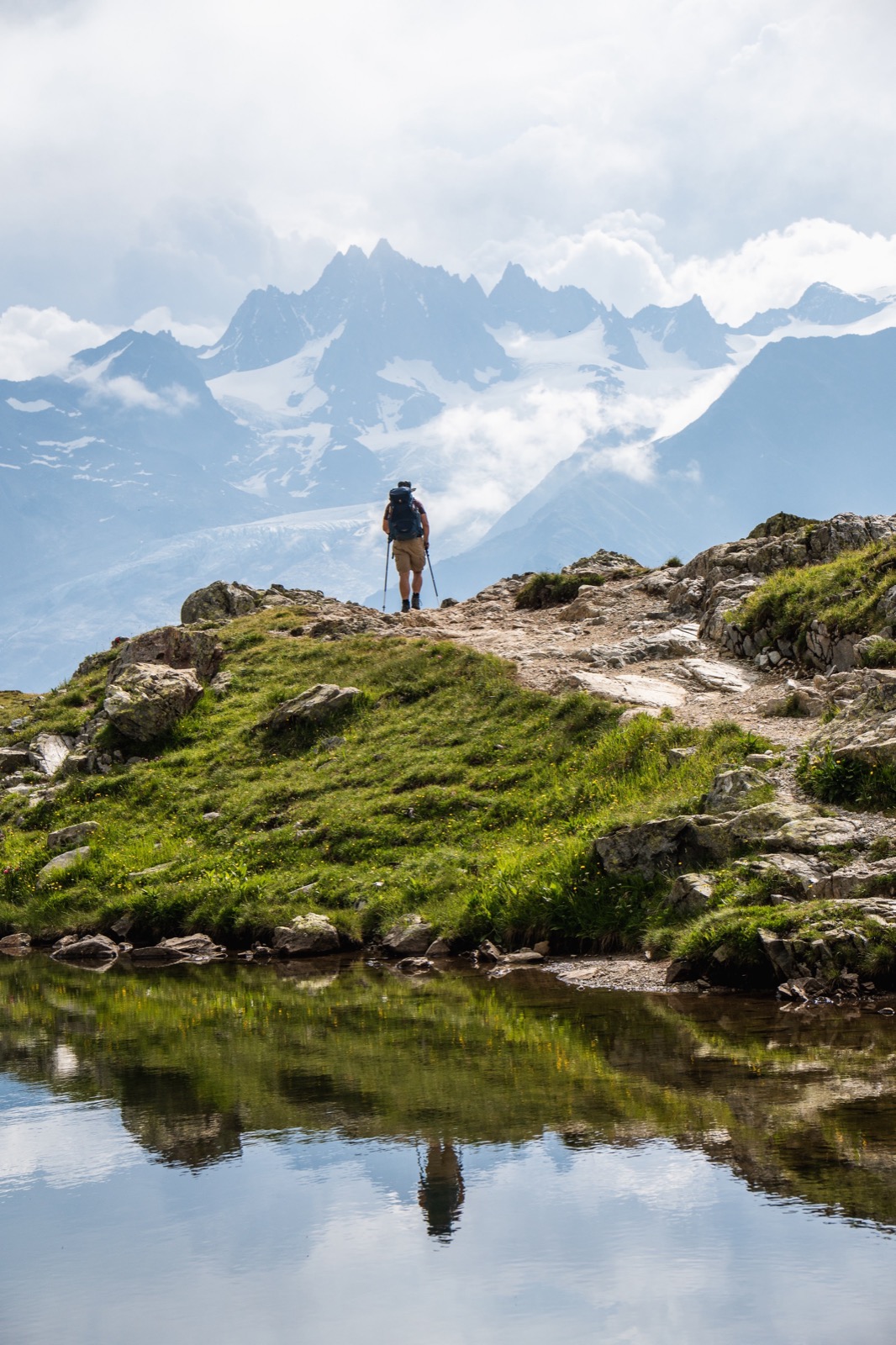 Runner standing on a rocky knoll, reflected in an alpine lake below snowy peaks